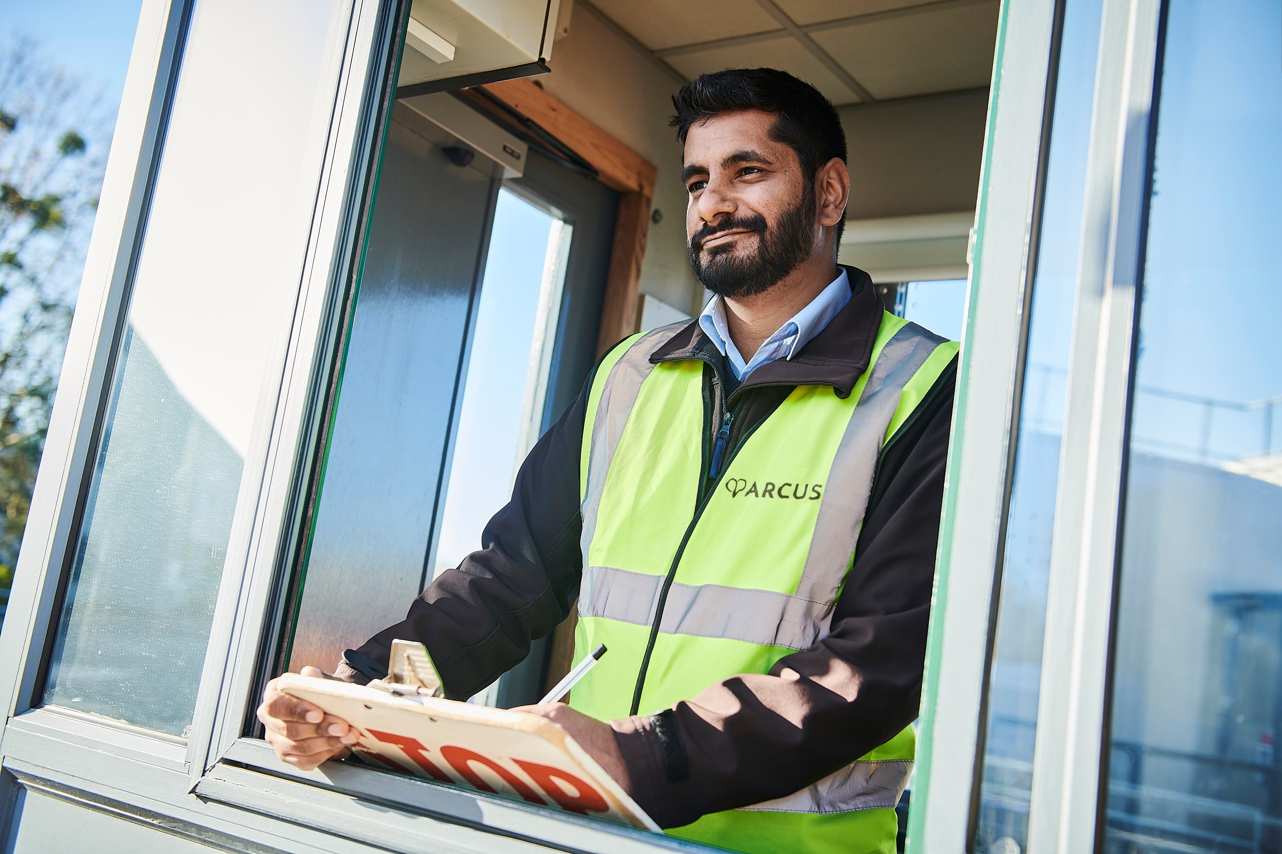 A man wearing a high-visibility vest is standing in a small booth, holding a clipboard. He appears to be working as a security or gatehouse officer, likely monitoring or checking vehicles or personnel. The booth has large windows, allowing a clear view outside on what seems to be a bright day. The man is looking out with a calm expression, with natural light reflecting off the glass around him. The scene suggests a professional work environment, possibly at a business or industrial site.