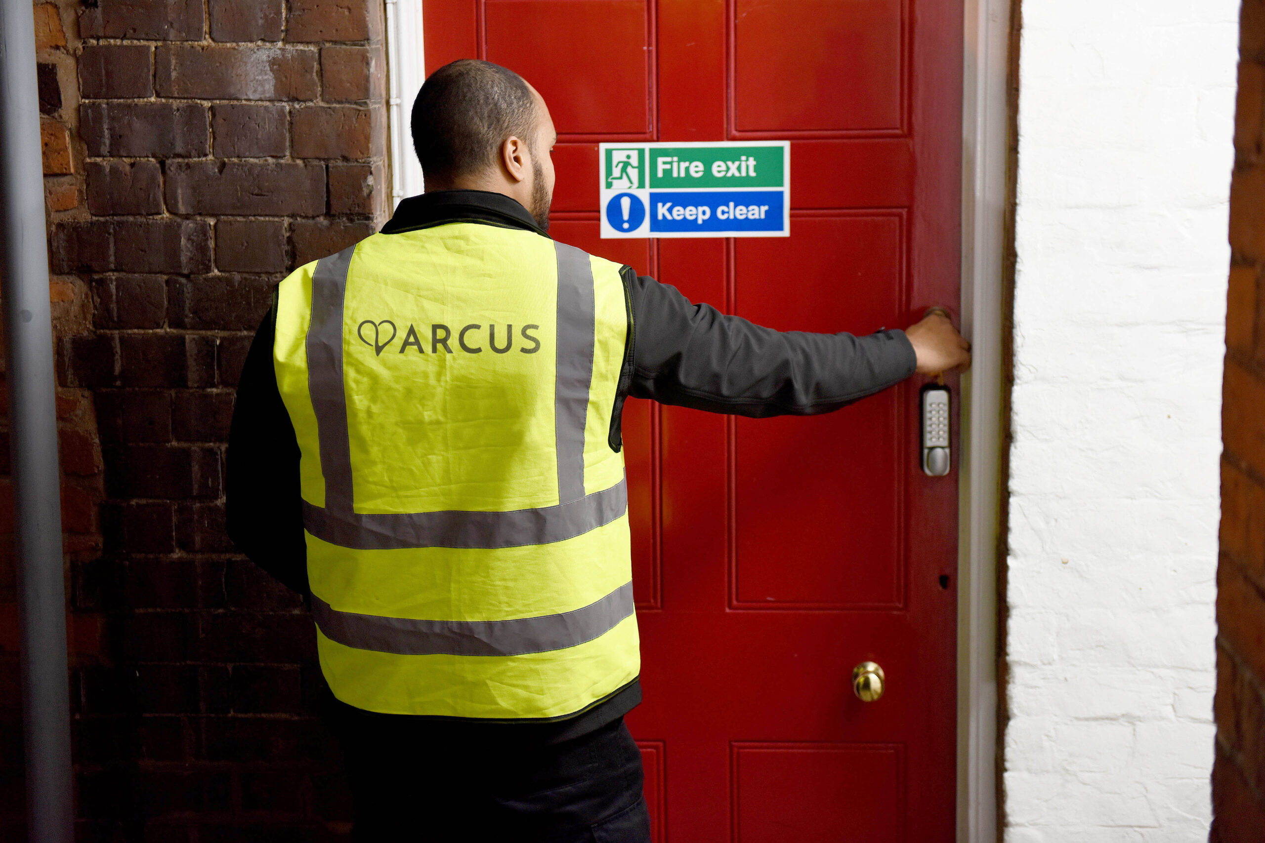 A man in a high-visibility vest is seen from behind as he opens a red door using a keypad entry system. The door is marked with a fire exit sign that reads "Fire exit" and "Keep clear." The man is positioned in a corridor with brick walls, indicating he may be performing security or safety-related tasks. The scene suggests the worker is either entering or securing an area, ensuring compliance with safety regulations.