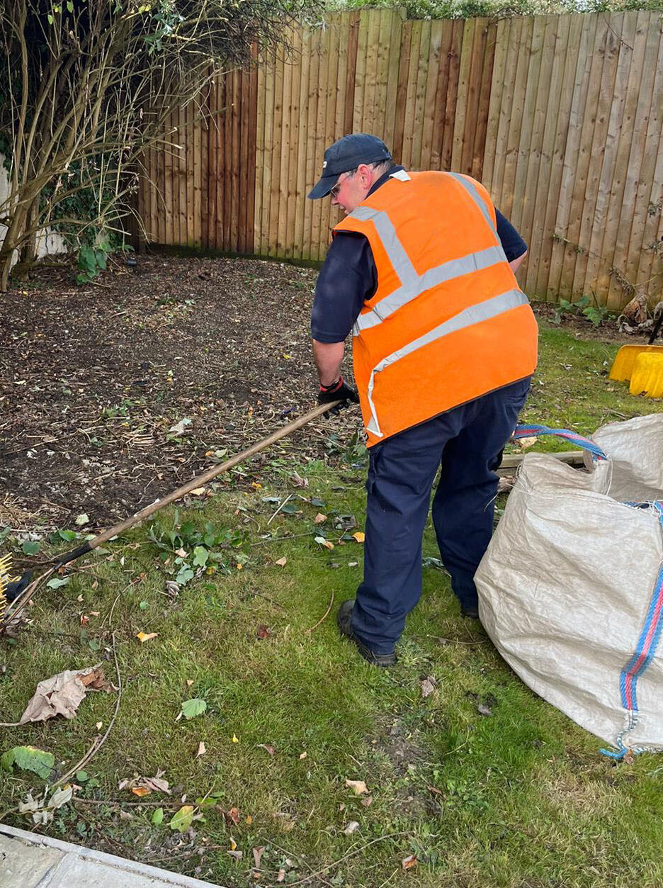 A worker in a high-visibility orange vest and dark trousers is clearing vegetation in a garden area. The worker is using a rake to gather leaves, branches, and debris, which are spread across a patch of bare soil and grass. A large white sack for collecting the waste is placed nearby. The garden is enclosed by wooden fencing, and a large bush with bare branches extends over the area being cleared. The scene suggests routine garden maintenance, focused on tidying and removing overgrown vegetation.