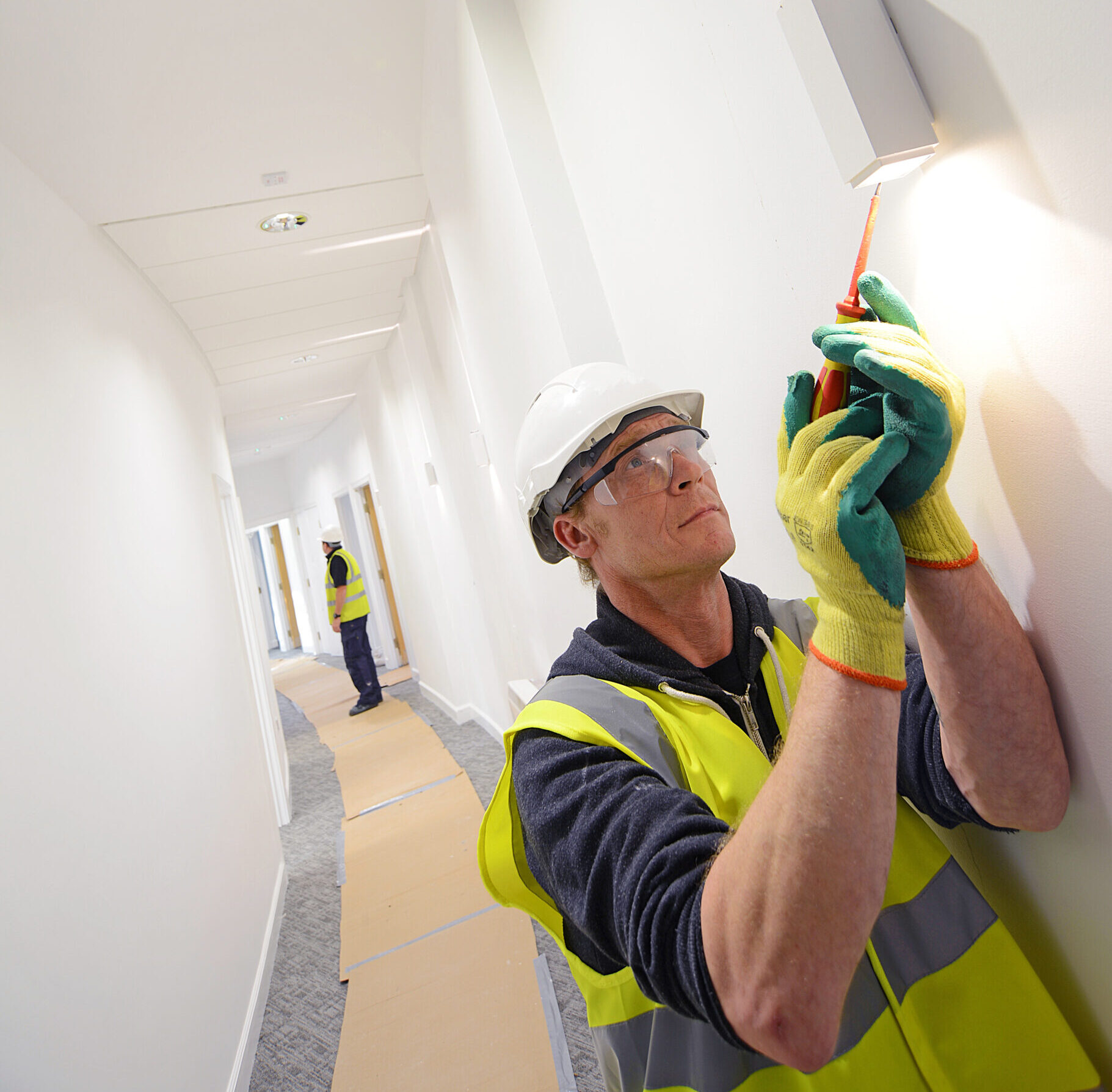 A worker in a high-visibility vest, hard hat, safety glasses, and protective gloves is focused on adjusting or installing a wall-mounted light fixture in a well-lit hallway. He carefully uses a screwdriver to work on the fixture. In the background, another worker in similar safety gear stands further down the corridor, which has been protected with floor coverings, likely to prevent dust and damage during maintenance. The scene conveys a sense of precision and attention to safety in a professional maintenance or installation environment.