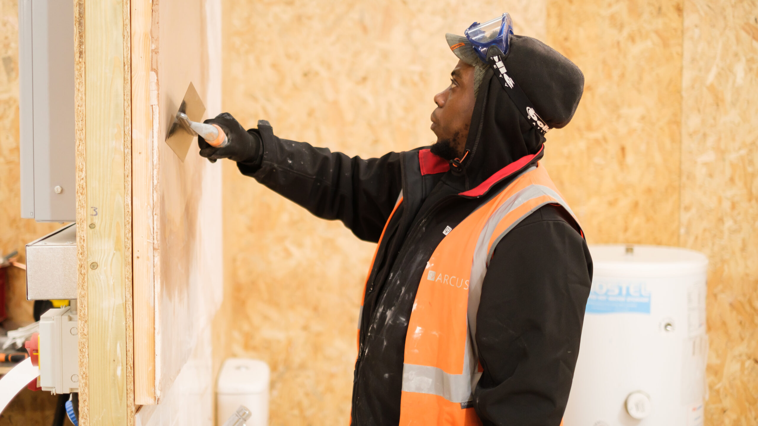 A worker in an Arcus-branded high-visibility vest is applying plaster to a wall using a trowel. The individual is focused on the task, wearing a black jacket and a beanie with protective goggles resting on top. The background consists of a construction or training area with wooden wall panels and equipment, indicating a setting for practical hands-on work or training in a construction skill. The worker’s careful attention to detail highlights the precision required in plastering.