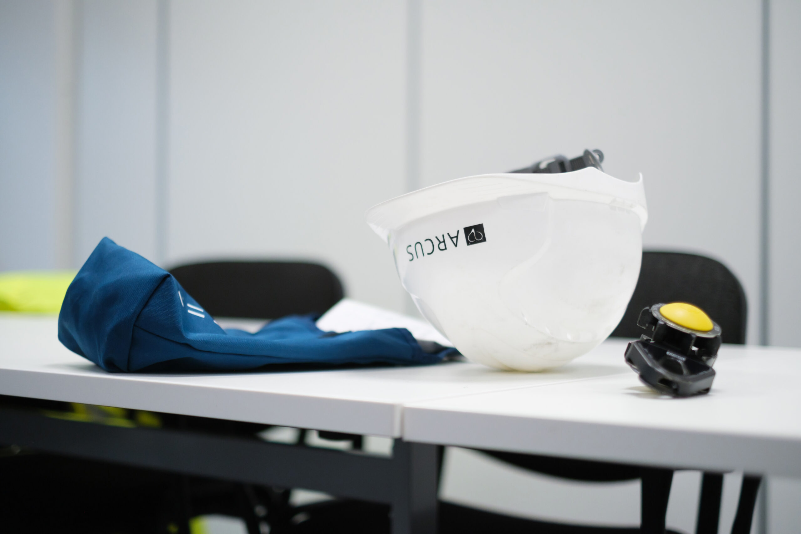 A white safety helmet branded with "Arcus" rests on a table in a training or meeting room. Next to the helmet lies a folded piece of blue protective clothing and a black and yellow safety device. The background features simple, blurred office furniture, emphasising the safety equipment in the foreground. The image suggests preparation for a safety briefing or a work session where protective gear is essential.