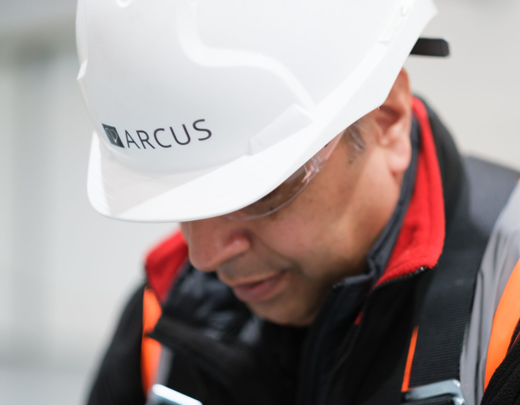  A close-up image of a worker wearing a white safety helmet branded with "Arcus" and a high-visibility jacket. The worker appears focused, looking downward while engaged in a task. Safety glasses are worn for eye protection, and the worker's clothing includes a red inner layer for warmth. The blurred background emphasises the worker's concentration and commitment to safety procedures, highlighting the Arcus brand's dedication to professionalism and safety.